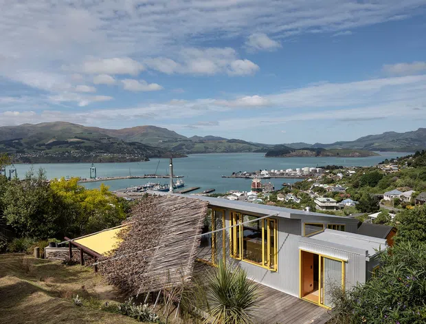 Elevated exterior view of a modern low-rise dwelling overlooking a sheltered coastal bay and surrounding hills. The building has corrugated grey cladding, extensive yellow-framed glazing and doors, a flat or shallow-pitched roof, and a timber deck. A thatched-roof outdoor structure sits adjacent to the house, with marina buildings and waterfront settlement visible beyond.
