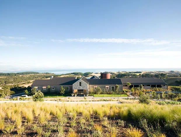 A wide exterior view of a low, sprawling rural-style residence set within a grassy coastal or dune landscape. The building has dark pitched roofs, weathered grey cladding, stone-like central masonry, and multiple connected wings. Open countryside and a broad horizon extend behind the house.