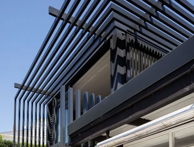 Upward-looking close view of a contemporary building exterior with bold black steel framing, projecting horizontal beams, and vertical fins forming a geometric layered facade. White masonry walls, glazed windows, and a terrace or balcony structure are visible beneath the open framing, set against a clear blue sky and landscaped residential surroundings.
