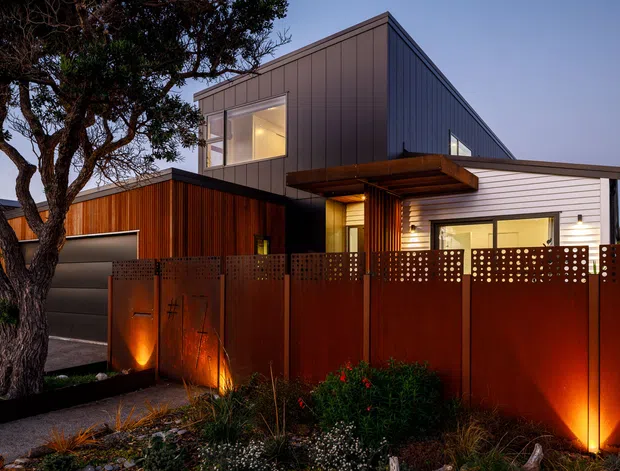 Contemporary two-storey residence photographed at dusk, featuring a strong geometric form with a dark grey standing-seam or panelled upper volume, white horizontal cladding, and warm timber-clad sections around the entry and garage. A rust-toned perforated metal privacy fence encloses the front garden, with integrated landscape lighting, native-style planting, and a mature tree in the foreground. Large windows and a projecting flat-roofed entry canopy contribute to the modern architectural character.
