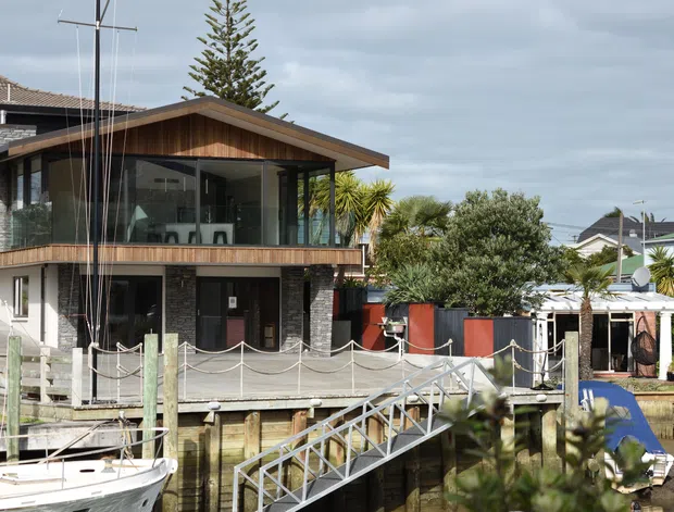Exterior view of a contemporary multi-level waterfront home with a prominent timber-clad upper pavilion, extensive dark-framed glazing, exposed balcony, and a gabled roof. A marina-style timber deck, boat ramp, and dock occupy the foreground, with neighboring homes and coastal vegetation surrounding the property.