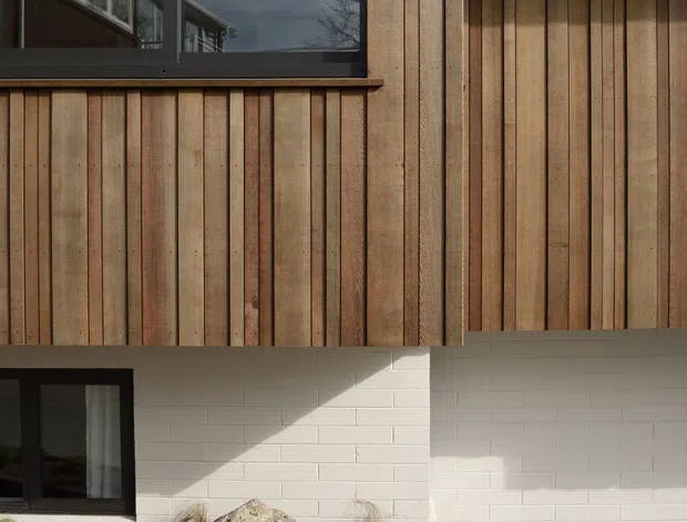 Close-up exterior view of a contemporary building facade combining vertical natural timber cladding with white-painted masonry at ground level. Dark-framed windows reflect the surrounding structure and sky. A simple concrete path, gravel planting bed, grasses, and a large boulder sit along the base of the building.