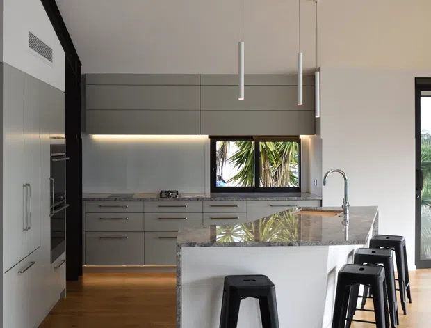 A contemporary open-plan kitchen with a large white island, waterfall stone benchtop, integrated grey cabinetry, stainless-steel appliances, and black bar stools. Warm timber flooring contrasts with the cool grey joinery. Linear pendant lights, under-cabinet lighting, a skylight, and large glazed openings create a bright, refined space.