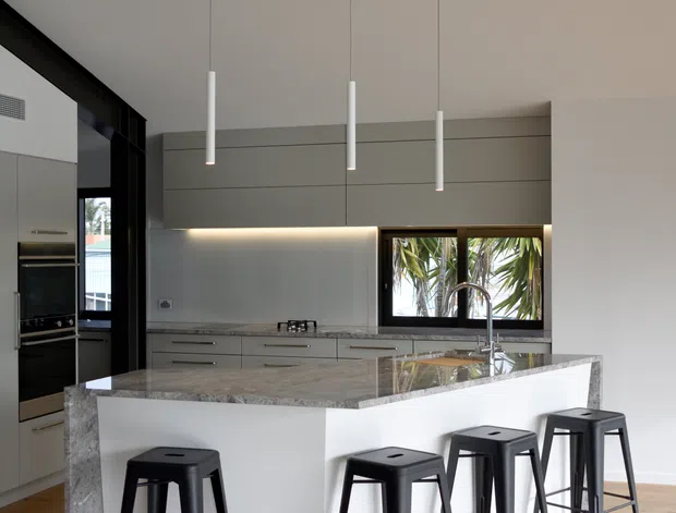 Contemporary kitchen with a large white waterfall island, polished stone benchtop, integrated light-grey cabinetry, dark-framed glazing, and three slim white pendant lights. Black bar stools provide contrast, while tropical foliage is visible through the window.