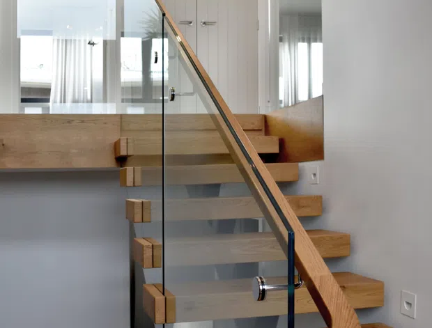 Interior view of a contemporary staircase with floating timber treads, a sculptural curved timber handrail, and clear glass balustrades supported by dark metal fittings. The stair rises through a bright hallway or landing, with white walls, mirrored surfaces, and white paneled doors visible around it.