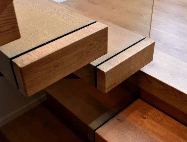 Close-up interior view of a modern timber staircase with thick oak-toned treads and integrated stair stringers, accented by slim black reveals. A frameless glass balustrade runs alongside the stairs. The landing has matching timber flooring, a vivid red flush door, white skirting, and a black-framed window.