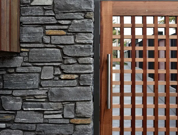 Close-up architectural detail of a contemporary entrance, showing rough stacked dark stone wall cladding beside a warm timber-framed glazed door with a square timber lattice screen and vertical metal pull handle. A courtyard or exterior area is visible beyond the door.