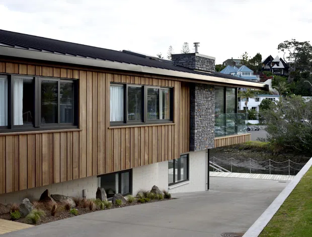 Exterior architectural view of a contemporary multi-level house on a sloped site. The building features vertical timber cladding, a dark pitched metal roof, large dark-framed windows, a stone-clad chimney or feature wall, and a pale rendered lower level. A driveway and landscaped foreground are visible, with neighbouring hillside houses in the background.