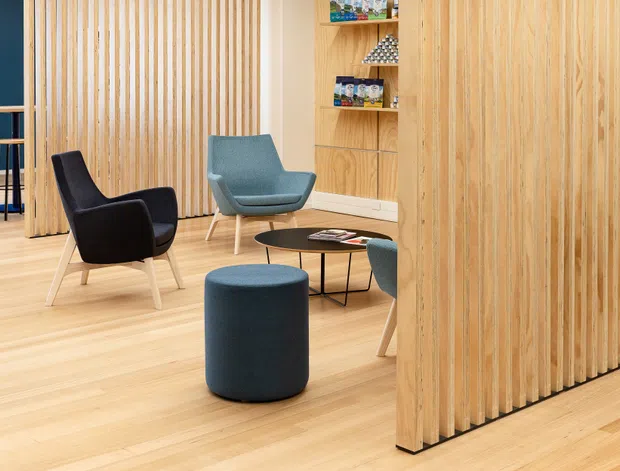 A contemporary commercial lounge or waiting area featuring pale timber flooring and extensive vertical timber slat wall screens. A black lounge chair, muted blue lounge chair, round blue ottoman, and low round coffee table form a seating arrangement. Open shelving displays small items, while a bar-height seating area is visible beyond the slatted partition.