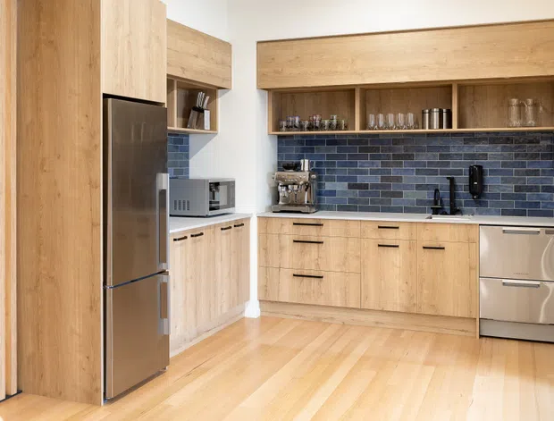Bright contemporary kitchen featuring extensive light timber cabinetry, integrated open shelving, a dark blue-grey brick-pattern tiled splashback, stainless-steel refrigerator and dishwasher, and pale timber flooring. Vertical timber slats and a compact appliance nook add architectural detail.