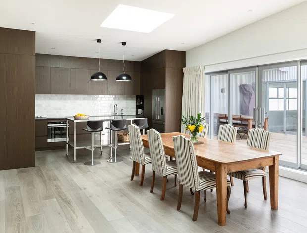 Bright open-plan kitchen and dining area with dark timber cabinetry, a stone-look backsplash, central island with bar seating, and a large timber dining table surrounded by upholstered chairs. Full-height sliding glass doors open to a deck, while a roof skylight and pendant lights provide additional illumination.