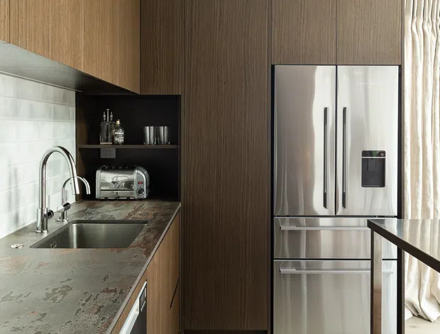 A compact contemporary kitchen with tall dark timber-grain cabinetry, integrated storage, a stainless-steel refrigerator, and a stone-look countertop with an undermount sink and brushed metal tapware. A white tiled splashback, open shelf with small appliances, pale timber flooring, and a narrow timber-framed island or table complete the space. Natural light enters through curtains at the right.