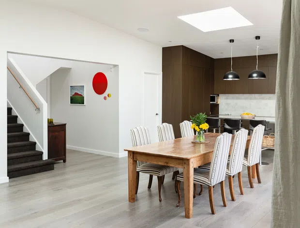 Bright open-plan dining and kitchen interior with a large timber dining table surrounded by upholstered chairs. The space features pale timber-look flooring, white walls, a dark timber kitchen with a light stone splashback, black pendant lights, a skylight, and an adjacent carpeted staircase. Contemporary furnishings include colourful wall art and a floral table arrangement.