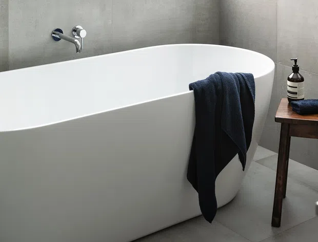A minimalist contemporary bathroom featuring a freestanding white oval bathtub against large grey concrete-look wall tiles. A chrome wall-mounted tap, dark towel, and a simple dark timber side table with soap dispenser add contrast. Part of a white vanity is visible at the right edge.