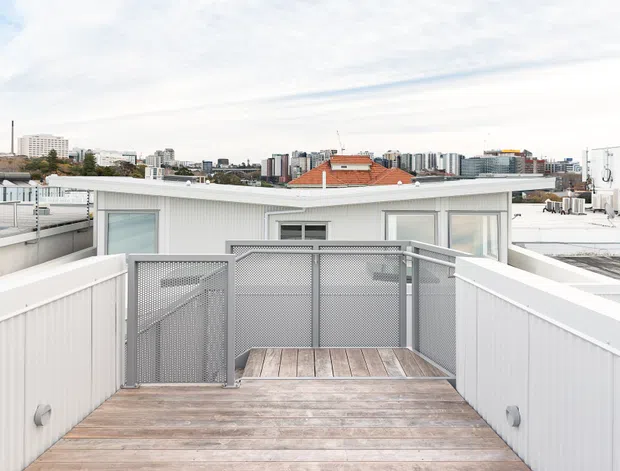 A rooftop terrace or deck with weathered timber decking, white corrugated parapet walls, and grey perforated metal privacy screening and gate. Low modern rooftop structures and rooftop equipment surround the space, with an urban skyline visible beyond. The setting is bright and contemporary with a restrained neutral palette.