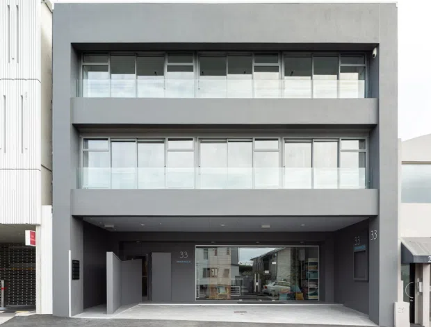 Street-level exterior photograph of a contemporary multi-storey residential or mixed-use building with a dark grey rendered facade, three stacked levels, broad recessed balconies, extensive glazed openings, and a covered ground-floor entry or garage-like frontage. The design has clean rectilinear lines and aluminium-framed glazing.