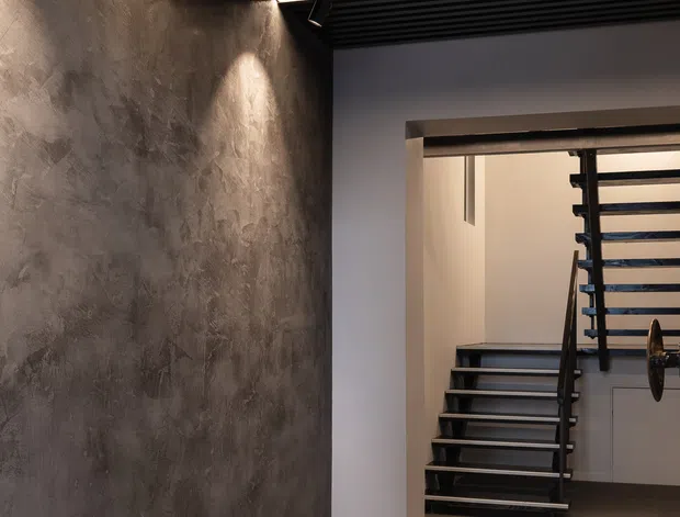 A moody modern interior hallway leading to an open-riser staircase. The space features a dark textured feature wall, large-format beige floor tiles, white walls, and a black slatted ceiling with integrated linear lighting. A sculptural black stair with open treads and a dark handrail is visible beyond the doorway.