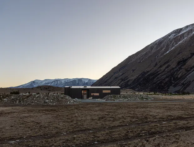 A low, dark-clad single-storey building sits in a stark alpine valley beneath a steep, rugged mountain slope. The broad gravelly foreground and sparse terrain create a remote, austere setting, while snow-dusted ridges and cool evening light reinforce the mountainous context.