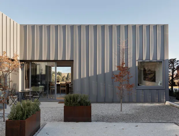 Modern single-storey building exterior with a sculptural vertically ribbed grey metal facade, large glazed openings, and a minimalist gravel courtyard. Rusted steel planter boxes with sparse landscaping sit in the foreground, while the clean geometric form and mountain landscape beyond reinforce the contemporary architectural character.