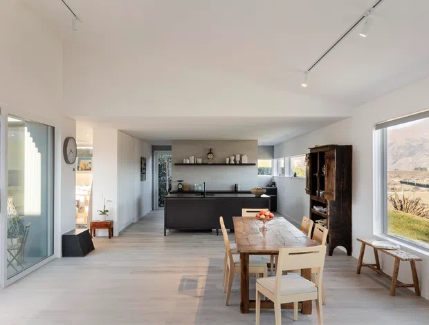Bright open-plan interior combining a kitchen, dining area and living space. The room has white walls and ceiling, pale timber flooring, a dark kitchen island with integrated cabinetry, a long dining table with timber chairs, and large glazed doors and windows overlooking the landscape. Track lighting and simple contemporary furnishings create an airy, calm atmosphere.