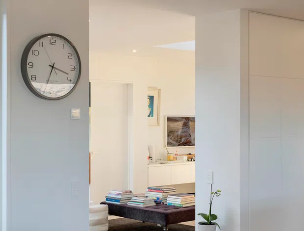 Bright contemporary residential interior showing a minimalist hallway or entry area opening into a living room. The space has pale timber flooring, white walls and ceiling, recessed downlights, extensive flush built-in cabinetry, a large wall clock, a low dark coffee table stacked with books, and a small timber side table with a potted orchid. The living area and artwork are visible beyond the opening.