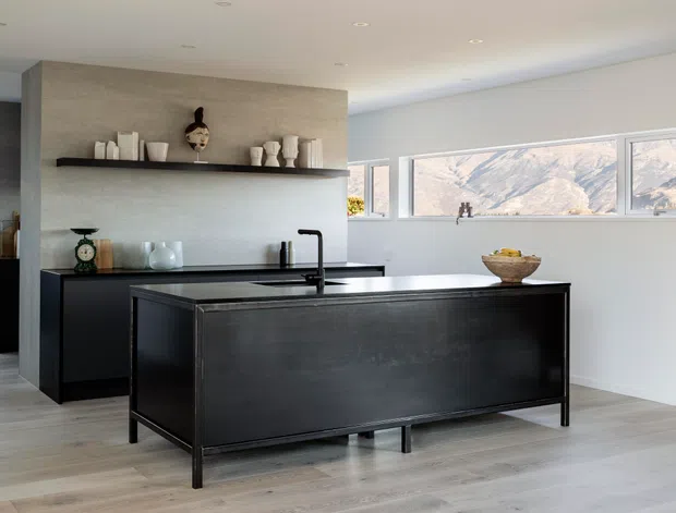A bright contemporary kitchen with a large black island, dark fixed cabinetry, pale timber-look flooring, and a textured grey feature wall. Open shelving displays neutral ceramic vessels, while wide horizontal windows provide a mountain view. The room has clean lines, recessed ceiling lights, and a restrained monochromatic palette.