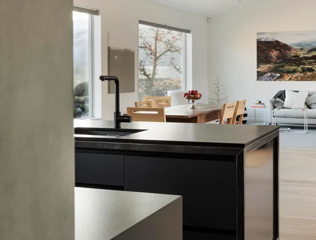 A bright, open-plan contemporary kitchen viewed from behind a partial wall, featuring a large matte-black island with an integrated sink and tall black tap. The kitchen opens to a dining area with a timber dining table and chairs and a light-coloured living room beyond. Pale timber flooring, white walls, large windows, a skylight, track lighting, and mountain artwork create an airy, minimal interior.