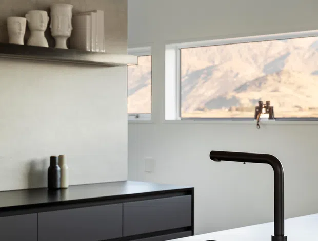 A minimalist contemporary kitchen focused on a dark matte island benchtop with an integrated black undermount sink and tall black gooseneck tap. Dark flat-panel cabinetry lines the rear wall, beneath a concrete-look feature wall and open shelf displaying sculptural ceramic vessels. Large horizontal windows bring in natural light and frame a rocky landscape.