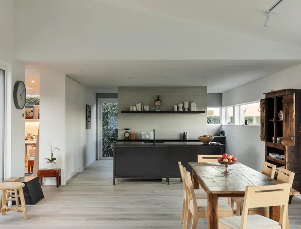 A bright open-plan kitchen and dining area with a contemporary dark kitchen island, pale timber flooring, white walls, and broad windows. A long timber dining table with light wood chairs occupies the foreground, while a tall rustic timber display cabinet provides contrast against the clean-lined kitchen. A small pantry or utility area is visible through the opening on the left.