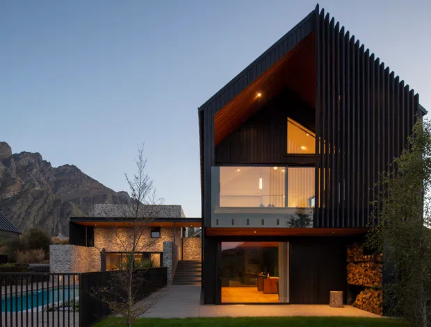 A dramatic modern multi-storey residence with a steep gabled form, dark vertical cladding and extensive glazing. Warm timber-lined soffits and illuminated interiors contrast with the dark exterior, while a brick-toned lower wing, stacked firewood and a fenced swimming pool sit in the foreground. Rugged mountain terrain forms the background.
