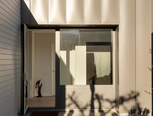 A contemporary residential entry facade viewed from a small gravel forecourt. The composition features a tall cream-coloured panelled exterior wall with strong vertical seams, a dark recessed entry canopy, a glazed front door and sidelight, and a horizontally clad side wall. Warm late-day sunlight creates deep shadows across the facade and concrete entry path.