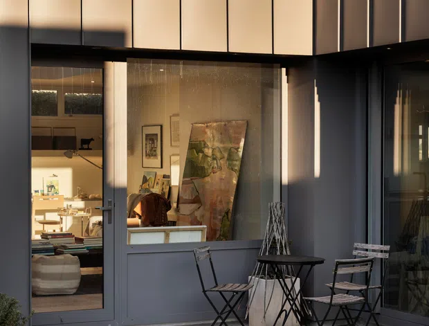 A compact modern courtyard or patio framed by dark metal and glazed doors beneath a tall vertical-panel facade. The space has a gravel ground surface, pale concrete paving, a small black metal bistro table and folding chairs, and a sculptural decorative feature. Interior living and dining areas are visible through the glazing, with artwork and warm lighting creating a connection between inside and outside.