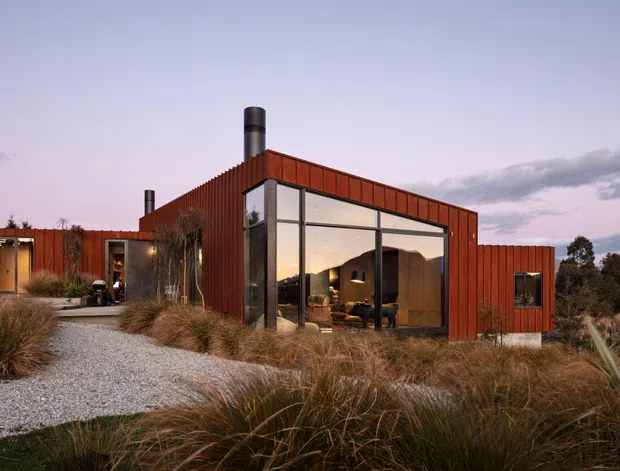 Single-storey contemporary home with a bold rust-red metal-clad exterior, large floor-to-ceiling glazed living area, and cylindrical metal chimney. The house sits within a landscaped garden of ornamental grasses, with a pale gravel path leading to the entrance. Warm interior lighting and timber furnishings are visible through the glazing at dusk.
