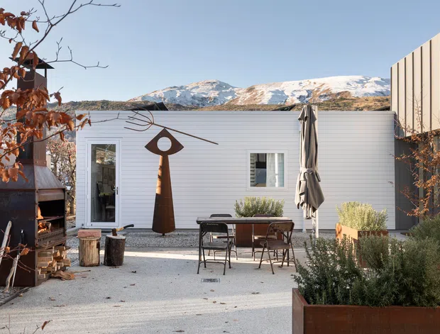 A contemporary outdoor courtyard terrace with pale gravel paving, a compact dining setting, closed umbrella, rusted steel planter boxes, sculptural artwork, and a built-in outdoor fireplace. A low white horizontally clad building and taller ribbed wall enclose the space, with snow-covered mountains visible beyond.