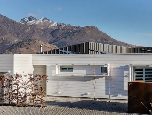 A low, contemporary single-storey building with pale horizontal cladding, crisp white window surrounds, dark louvred roof elements, and a sculptural dark entrance gate marked 99. The building sits in a minimalist gravel landscape, with rugged snow-dusted alpine mountains prominently visible behind it under a clear blue sky.