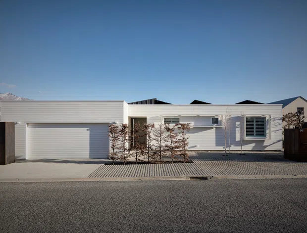 Street-facing view of a low, single-storey modern residence with a white horizontal-panel facade, integrated double garage, flat roof sections, broad windows with external louvres, timber privacy fences, and a patterned paved frontage. Snow-capped mountains are visible in the distant background.