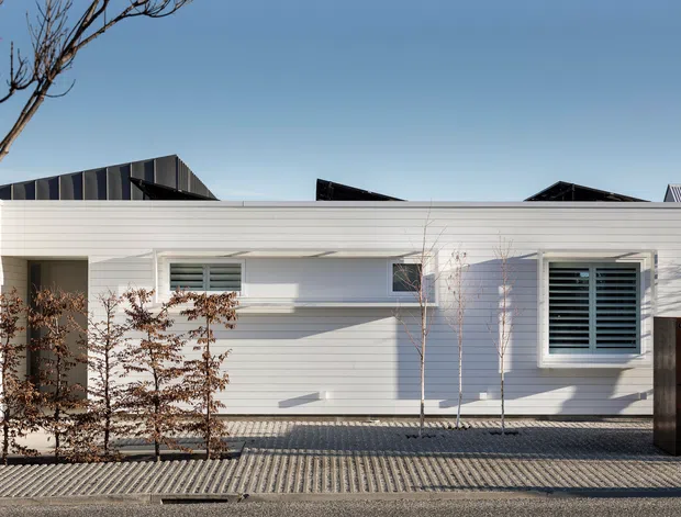 Street-facing view of a low, single-storey contemporary residence with a crisp white horizontal weatherboard facade, projecting flat and shallow pitched roof forms, deep-set openings, and prominent louvred windows. A paved forecourt and sparse planted screening sit in front of the house, with neighbouring rooflines visible behind.
