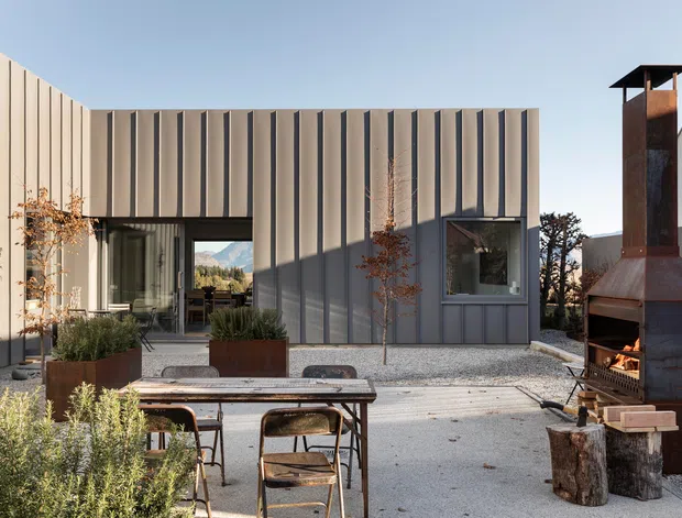 A contemporary courtyard terrace viewed from outdoors, featuring a vertically ribbed grey metal-clad building, large glazed openings, gravel paving, raised planter boxes, and an outdoor dining table with chairs. A tall rusted steel outdoor fireplace or chimney structure dominates the right side, with landscaped planting and mountain scenery visible through the opening.