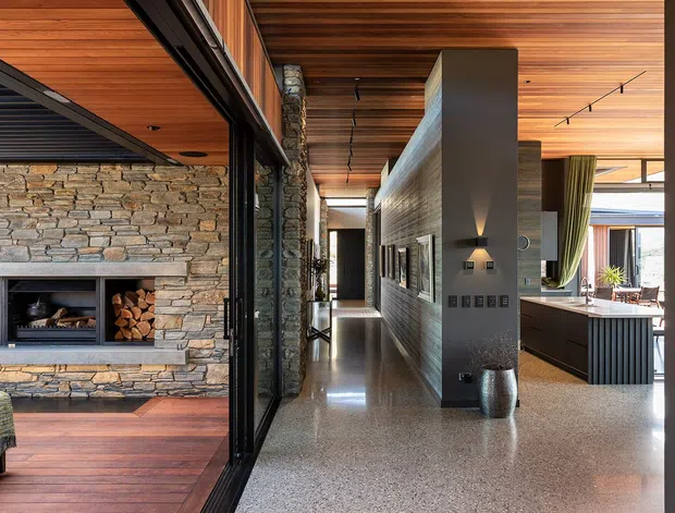 A refined open-plan residential interior with a long central hallway connecting a living area and kitchen. The living zone features a substantial stacked natural-stone fireplace and timber flooring, while the hallway and kitchen use polished terrazzo flooring, dark built-in joinery, and extensive warm timber ceiling lining. Black-framed glazed partitions create visual separation while maintaining openness. The kitchen includes a large island, and the space is lit by recessed and track lighting.