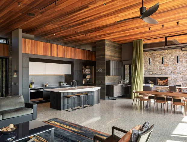 Open-plan contemporary living, kitchen and dining space with a dramatic exposed timber-lined ceiling, dark built-in cabinetry, a stone kitchen island with bar seating, and a dining area beside expansive glazing. A textured natural-stone fireplace wall forms a strong focal point, while polished terrazzo-like flooring, black-framed windows, and layered timber joinery create a warm, refined interior.