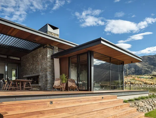 Contemporary mountain home exterior with broad timber decking and steps, a low-pitched projecting roof, extensive floor-to-ceiling glazing, dark metal-framed windows, warm timber soffits, and a prominent rough stone chimney and wall. An outdoor dining area and lounge chairs sit beneath the covered terrace, with rugged mountains visible beyond.