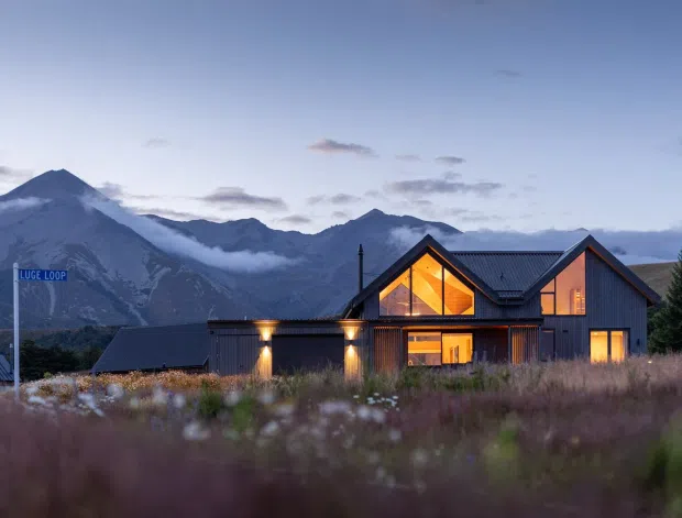 A contemporary dark-clad mountain lodge viewed at dusk across a grassy field, with warm interior lighting glowing through large windows and gable-end glazing. The building has multiple pitched gabled roof forms, timber-toned vertical cladding, and a dramatic alpine mountain backdrop.