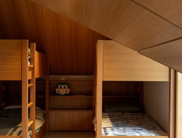 Compact children's bedroom with built-in wooden bunk beds, integrated shelving and cabinetry, warm wood wall and ceiling panelling, and a light carpeted floor. Strong natural light enters through the window on the right, creating a warm, cozy timber-lined interior.