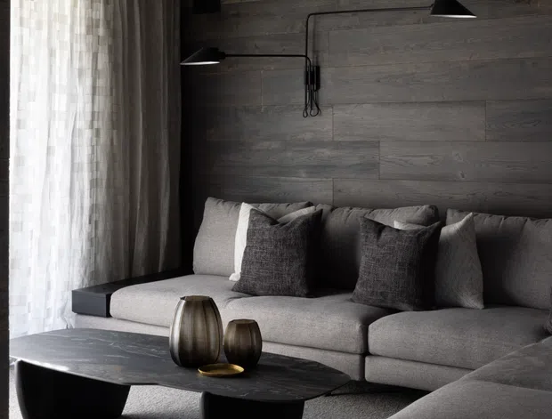 A moody, contemporary living room with dark horizontal timber wall and ceiling panelling, a large grey sectional sofa, black sculptural coffee tables, sheer floor-to-ceiling curtains, and minimalist black wall-mounted lamps. The room has a restrained monochromatic palette with warm natural timber tones and soft daylight.