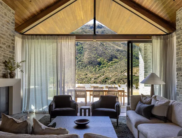A warm, light-filled living room with a dramatic pitched timber ceiling and exposed dark structural beams. Full-height glazing opens to a balcony or terrace with a mountainous landscape beyond. Rough pale stone walls frame a minimalist fireplace, while a large sectional sofa, leather lounge chairs, coffee table, and floor lamp create a comfortable seating area.