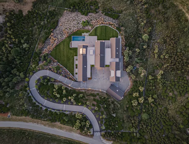 Aerial overhead view of a contemporary, elongated multi-wing residence set within a heavily landscaped, bush-like site. The building has several dark pitched or low-slope roof forms, pale rectangular roof sections, a small rectangular pool or water feature at one end, and a curved driveway winding through the planted grounds to the house. Dense native-looking vegetation surrounds the property.