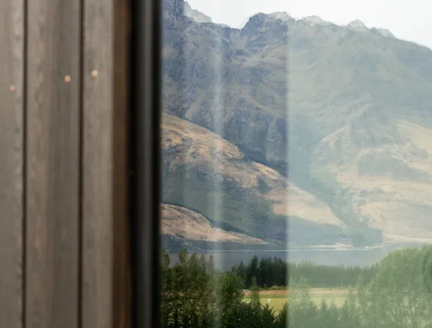 A vertical view through a large glazed opening, with dark timber wall cladding and a black window or door frame in the foreground. The glass looks toward a dramatic alpine mountain range, lake, tree-lined fields, and landscaped grounds. Reflections in the glazing add subtle interior and landscape overlays. The image is quiet and natural, with a strong connection between the building and its mountainous setting.