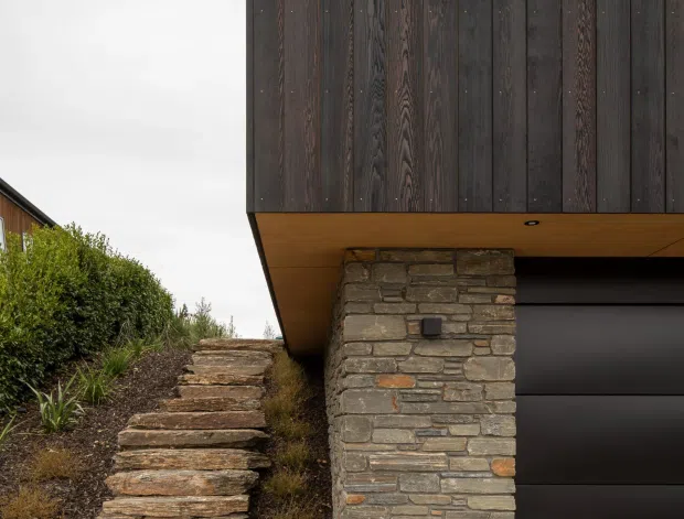 Close-up exterior view of a contemporary house entrance and garage. Dark vertical timber cladding forms the upper facade, supported by a stacked natural-stone pier. A black sectional garage door sits beneath a projecting timber-lined soffit, while rough stone steps rise through a landscaped slope alongside the building.