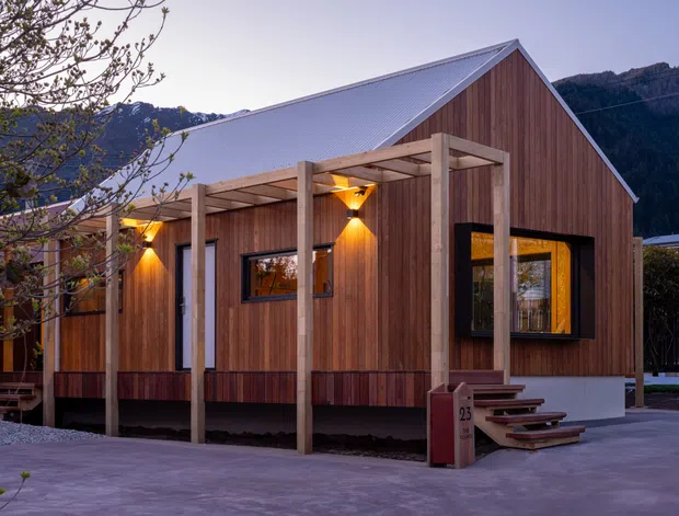 Exterior view of a compact single-storey gable-roofed building clad in vertical warm timber boards, with a pale corrugated metal roof, dark-framed windows, and a covered timber porch supported by simple posts. The building sits against a mountainous backdrop and is photographed in soft evening light.
