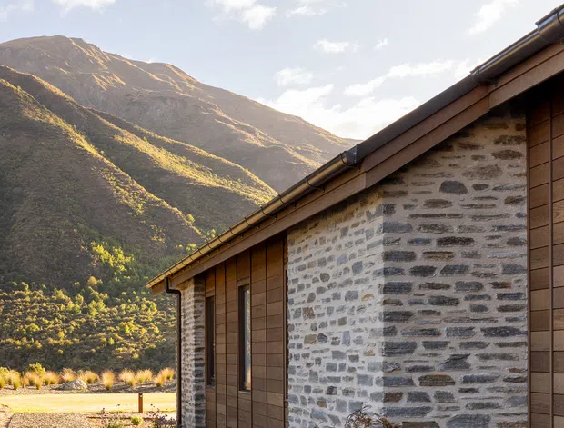 Exterior view of a low-slung mountain building with grey natural stone masonry walls, warm brown vertical timber cladding, dark metal gutters and a shallow pitched roof. A gravel path and sparse landscaped planting run alongside the facade, with a dramatic alpine mountain backdrop in warm late-day light.